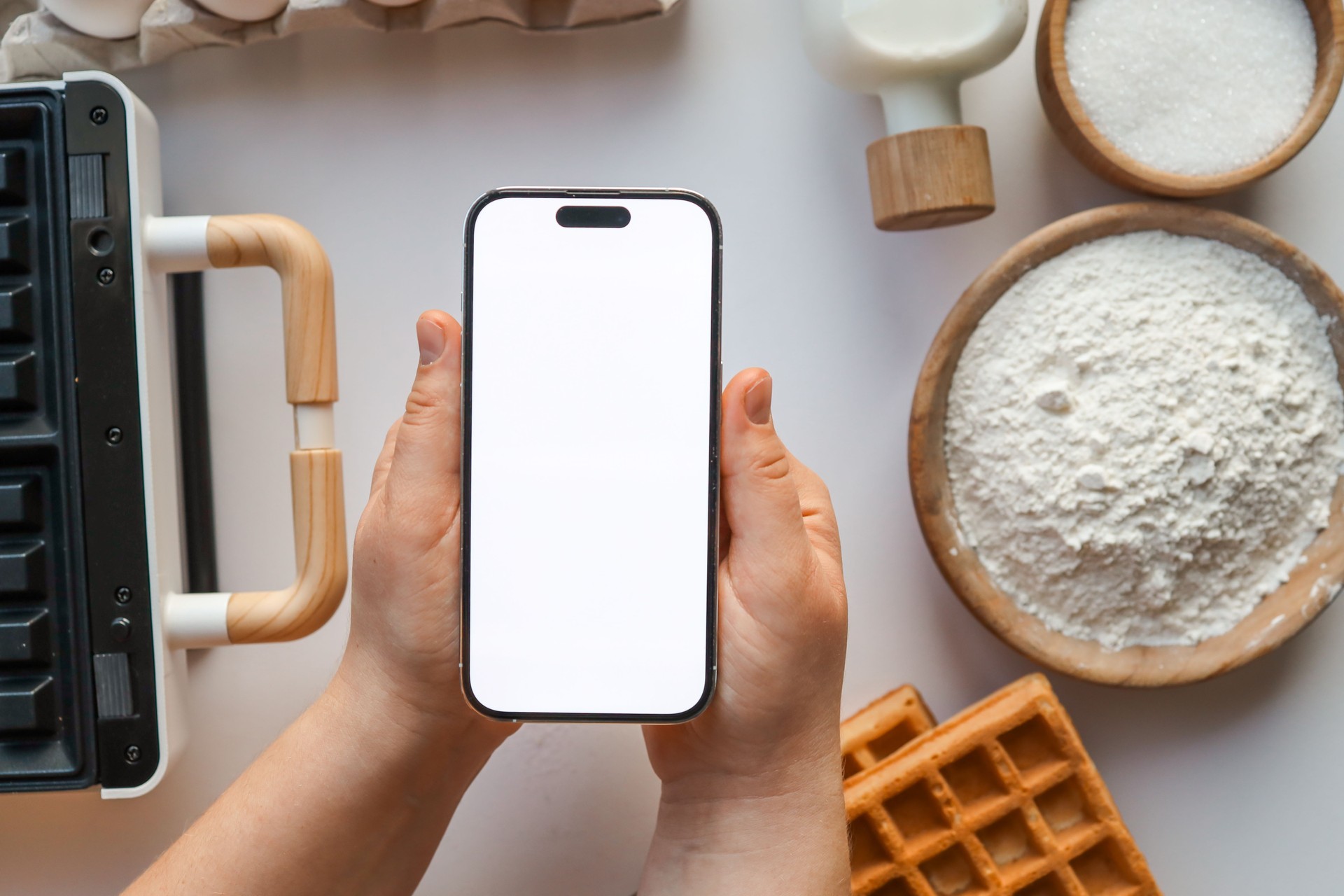 Hand holding a smartphone with a blank screen, surrounded by waffle-making ingredients including eggs, flour, milk, sugar, and a waffle iron on a white surface